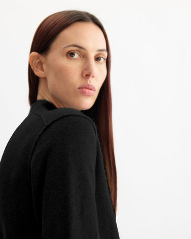 A person with long, straight brown hair and a neutral expression looks over their shoulder, wearing the Arlo Mott Perry Boxy Cardigan in Black against a plain white background.
