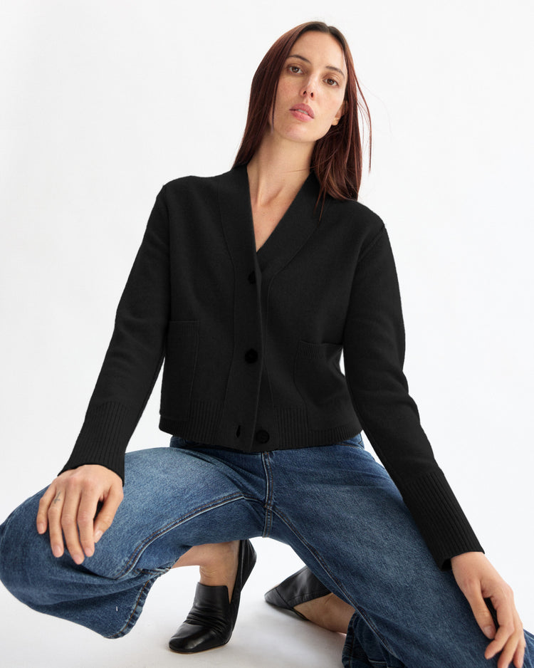 A person with long brown hair kneels on one knee, wearing the Arlo Mott Perry Boxy Cardigan in Black, blue jeans, and black shoes against a plain white background.